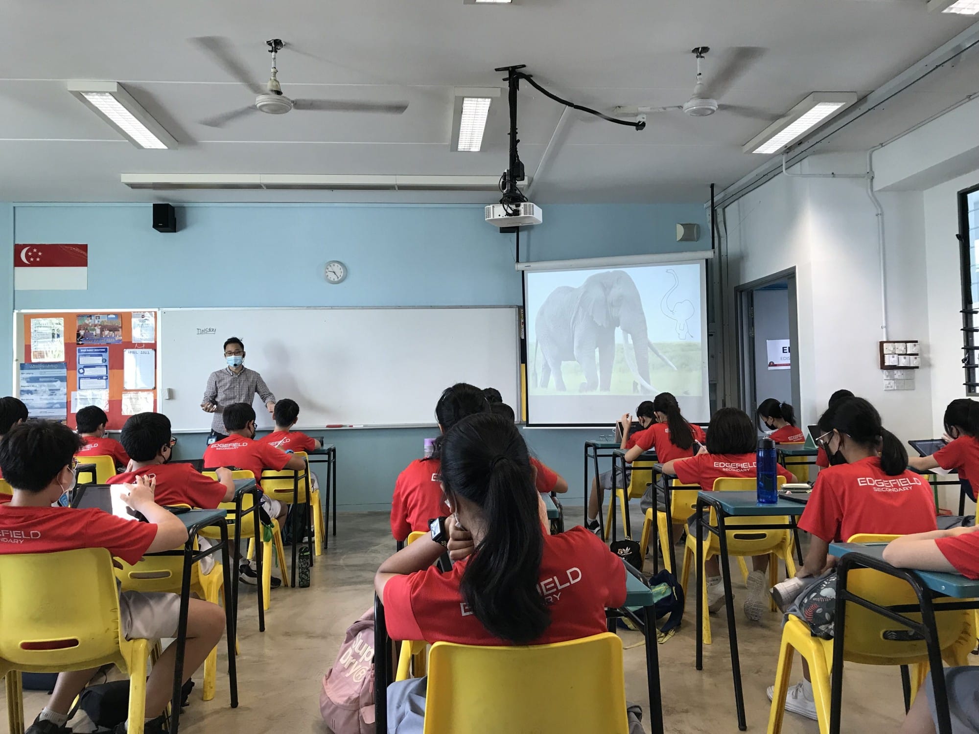 Students in red Edgefield Secondary shirts sit in a classroom watching a teacher present a slide showing an elephant.